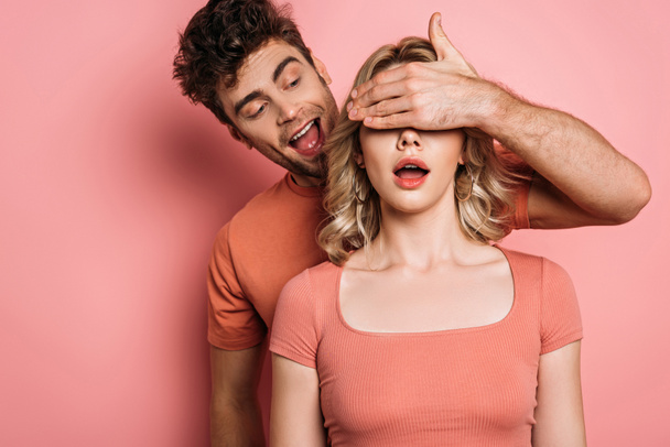 Хитрый мужчина закрывает глаза удивленной девушки с рукой на розовом фоне
 - Фото, изображение