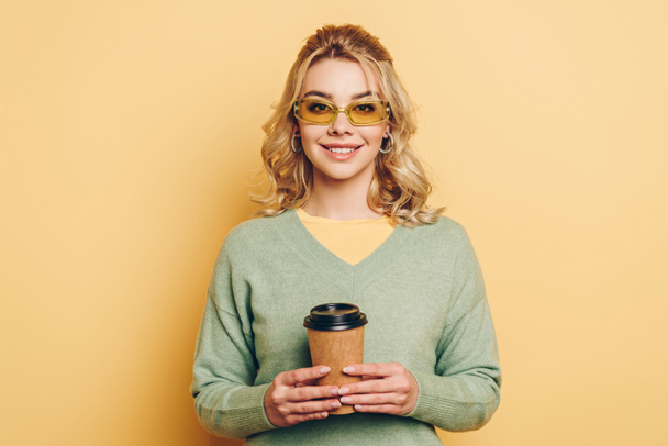 pretty, stylish girl holding coffee to go and smiling at camera on yellow background - Fotografie, Obrázek