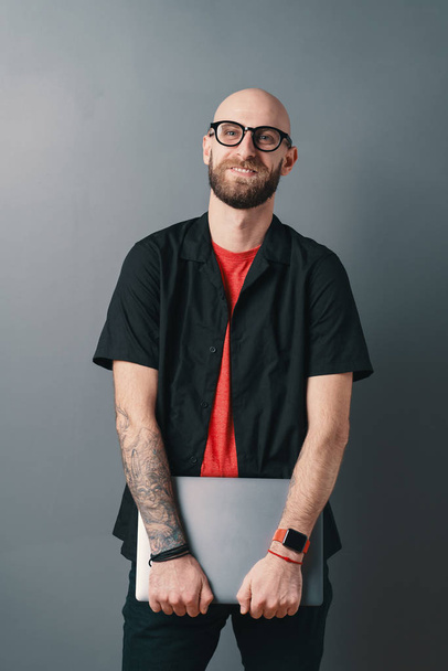 Χαμογελώντας νεαρός γενειοφόρος άνδρας με γυαλιά κρατώντας φορητό υπολογιστή κάτω από το χέρι στο στούντιο σε γκρι φόντο - Φωτογραφία, εικόνα