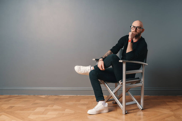 Um homem moderno sentado em uma pose confiante, perna sobre perna em uma cadeira, em fundo de parede cinza no estúdio
 - Foto, Imagem