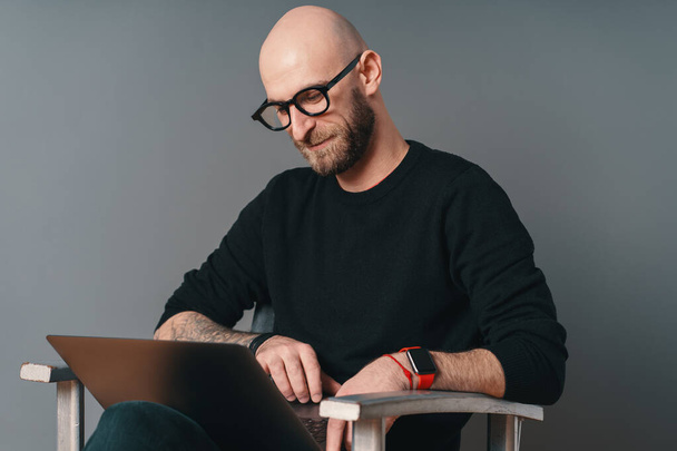 Άνθρωπος με γενειάδα και γυαλιά, γελώντας σερφάροντας στο διαδίκτυο σε ένα φορητό υπολογιστή Pc σε γκρι φόντο στούντιο - Φωτογραφία, εικόνα