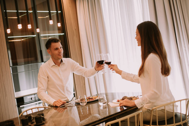 Sladký mladý pár s romantickou večeři a přípitek s sklenicemi červeného vína v luxusní kuchyni - Fotografie, Obrázek