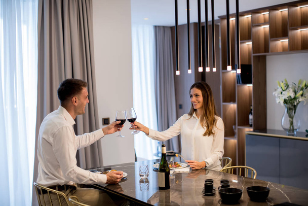 Sladký mladý pár s romantickou večeři a přípitek s sklenicemi červeného vína v luxusní kuchyni - Fotografie, Obrázek
