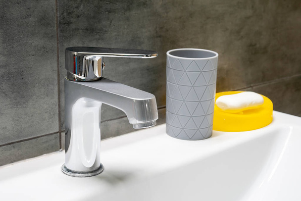 お湯と冷たい水のための溶存金属製の蛇口、歯ブラシ又は綿パッド用の灰色のカップ及び現代のバスルームの灰色のコンクリート壁に対する白いシンクに位置する黄色の石鹸皿 - 写真・画像