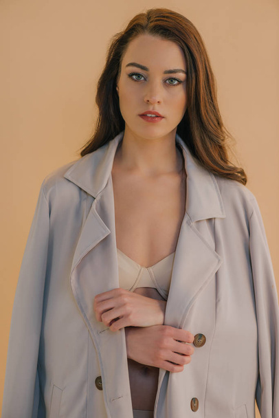 Beautiful young woman in coat and lingerie looking at camera isolated on beige  - Φωτογραφία, εικόνα