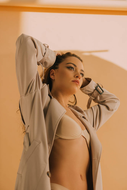 Low angle view of sexy girl in coat and underwear touching hair on beige background - Zdjęcie, obraz