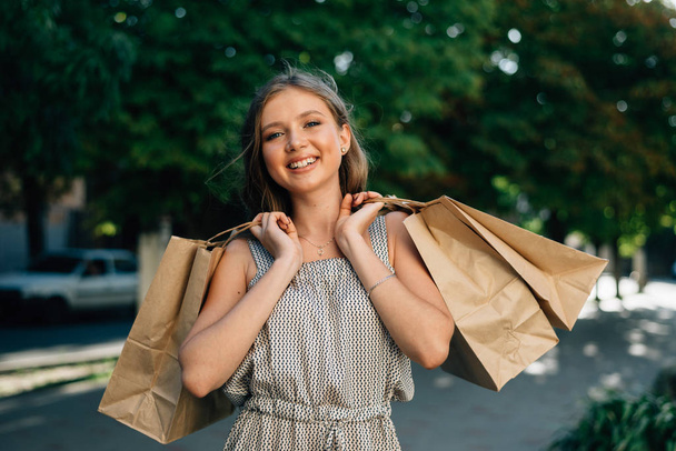 Egy gyönyörű nő csomagokat tart egy butikból mindkét kezében egy zöld utcán állva, közelről. - Fotó, kép