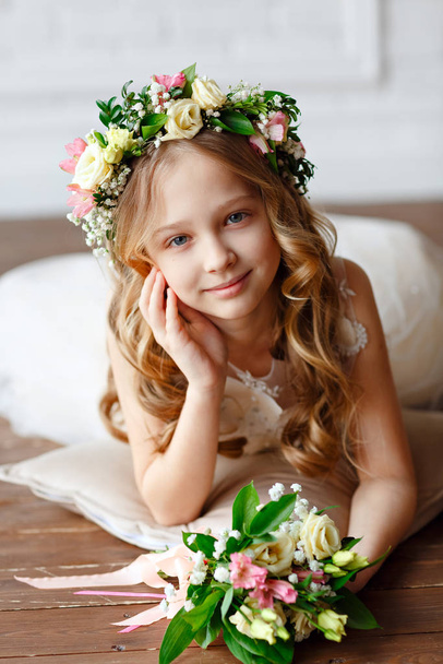 Kid - dziewczyna z blond włosami w koronie kwiat trzyma w rękach bukiet świeżych kwiatów. Piękna dziewczynka w jasnym studio leżąca na podłodze i spoglądająca w kamerę - Zdjęcie, obraz