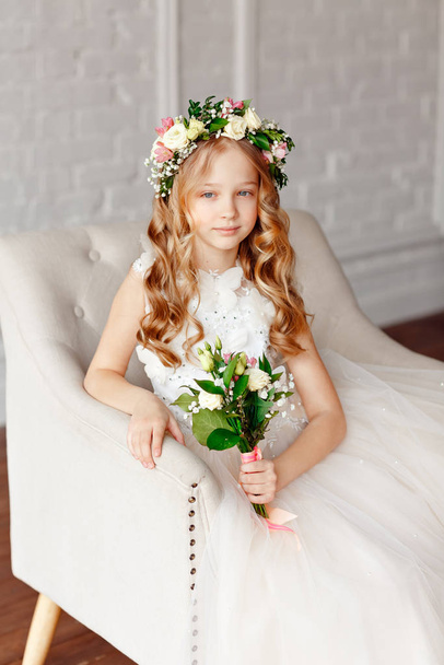 Beauty little girl angel with blond hair in a flower crown on her head holds in his hands a bouquet of fresh flowers and looks into the camera - Photo, image