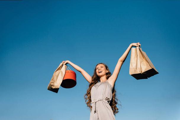 Hermosa mujer se regocija en las compras contra el cielo azul sosteniendo bolsas en dos manos
 - Foto, Imagen