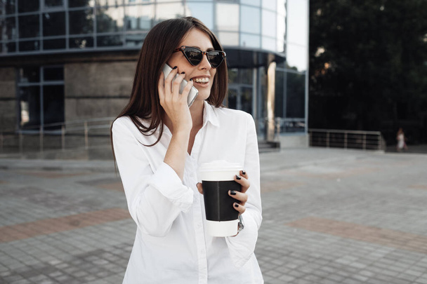 Portré egy divatos lány öltözött farmer és fehér ing iszik kávé és használja a Smartphone, Business Lady, nő Power Concept - Fotó, kép