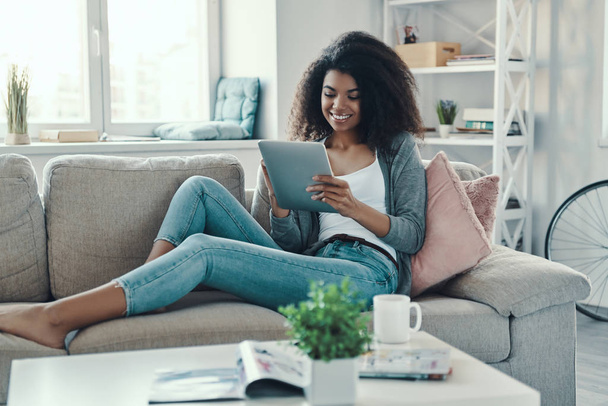 Relaxed young African woman using digital tablet and smiling while resting at home - Φωτογραφία, εικόνα