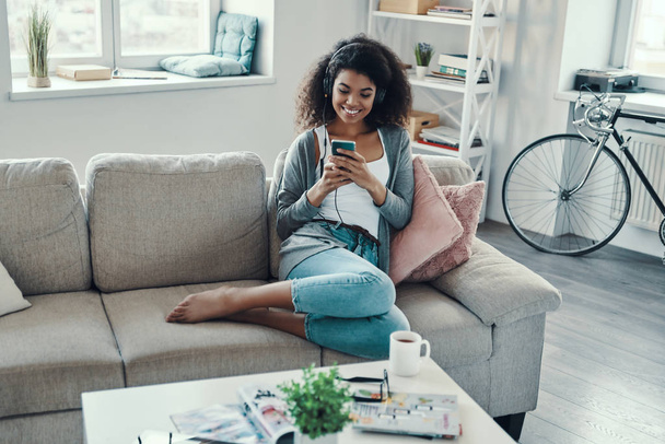 Beautiful young African woman in casual clothing using phone and smiling while resting at home                  - Zdjęcie, obraz