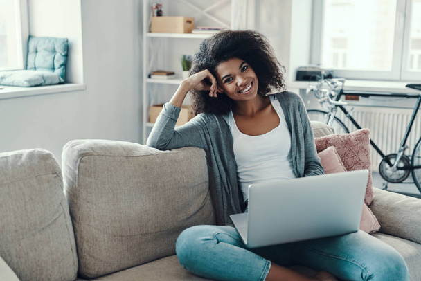 gelukkig jong Afrikaans vrouw in casual kleding met behulp van laptop en kijken naar camera terwijl rusten thuis                    - Foto, afbeelding
