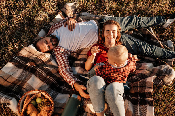 夏の週末のピクニックを楽しむ小さな息子と一緒に幸せな若い家族のお母さんとお父さん晴れた日の夕日、休暇時間のコンセプト - 写真・画像