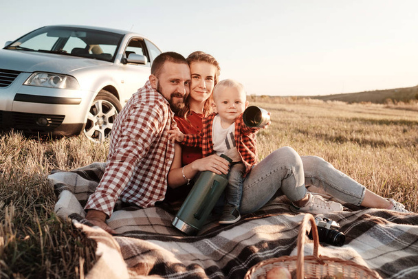 Onnellinen nuori perhe äiti ja isä pienen poikansa kanssa nauttimassa kesäviikonlopusta Picnic istuu Plaid Lähellä autoa kaupungin ulkopuolella pellolla Aurinkoinen päivä auringonlasku, loma ja road trip käsite
 - Valokuva, kuva