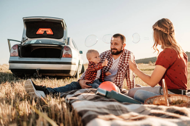 Feliz familia joven mamá y papá con su pequeño hijo disfrutando de un picnic de fin de semana de verano en el coche fuera de la ciudad, jugando con burbujas
 - Foto, Imagen