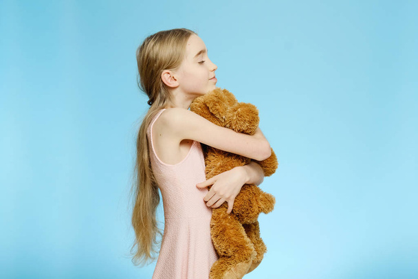 Το κορίτσι αγκαλιάζει ένα μεγάλο αρκουδάκι. Απομονωμένο πορτραίτο σε μπλε φόντο. Παιδική χαρά - Φωτογραφία, εικόνα