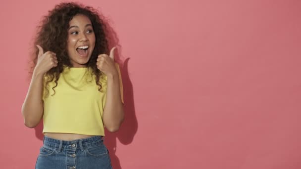 Joyful pretty curly woman showing thumbs up looking at the camera over pink background - Imágenes, Vídeo
