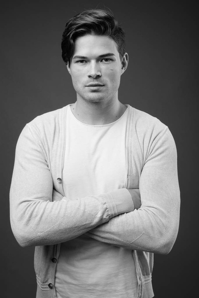 Studio shot of young handsome man wearing cardigan sweater against gray background in black and white - Zdjęcie, obraz
