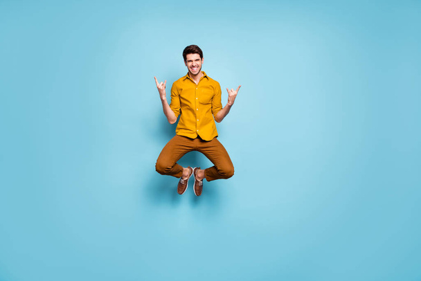 Full length photo of funny guy jumping high raising hands showing horns rejoicing rock concert wear yellow shirt pants isolated blue color background - Photo, image