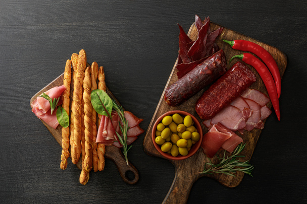top view of delicious meat platters served with olives, breadsticks and herbs on boards on wooden black table - Foto, imagen