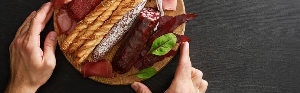 cropped view of man holding delicious meat platter served with breadsticks and herbs on board on wooden black table, panoramic shot - Photo, Image