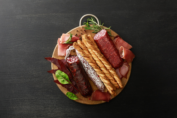 top view of delicious meat platter served with breadsticks and herbs on board on wooden black table - Photo, Image