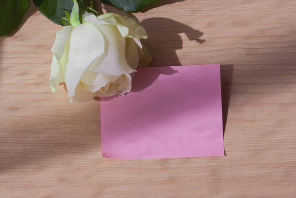 άδειο κενό ροζ χαρτί σημείωμα και λευκό τριαντάφυλλο στο τραπέζι από ξύλο με το φως του παραθύρου την ημέρα το πρωί - Φωτογραφία, εικόνα