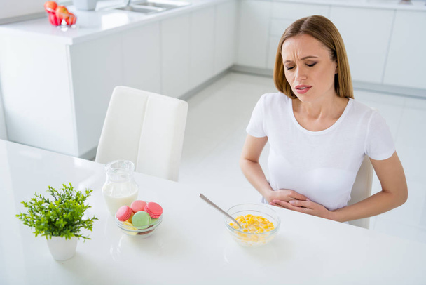 Photo of pretty upset housewife holding belly suffering terrible pain cant eat milk breakfast cornflakes have stomach upset sitting table white light kitchen indoors - Foto, immagini