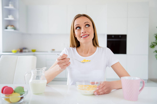 Photo of dreamer housewife holding spoon eating breakfast cornflakes healthy eating dieting looking dreamy up imagine herself chin sitting table white light kitchen indoors - Foto, afbeelding