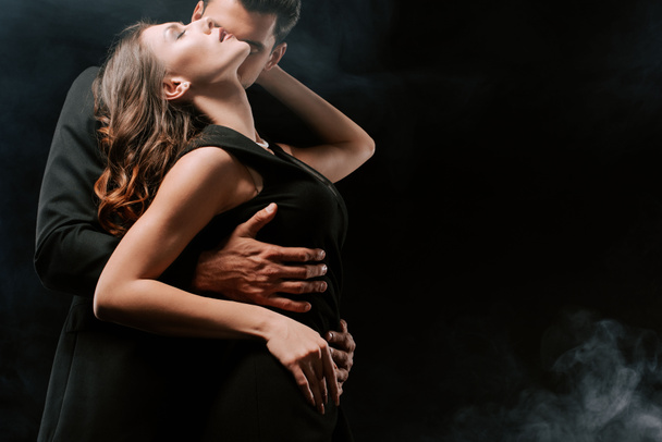 άντρας με κοστούμι αγκαλιάζει αισθησιακή γυναίκα σε μαύρο με καπνό  - Φωτογραφία, εικόνα