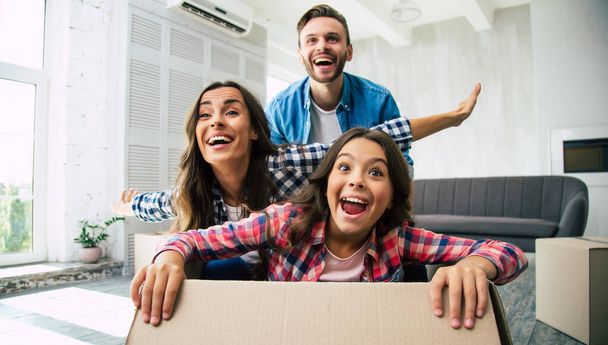 Father is playing with his family in their new home, mother and daughter are riding in a cardboard box laughing, all seem ecstatic and overfilled with positive emotions. - Foto, Bild