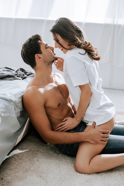 side view of cheerful girl touching handsome muscular boyfriend in bedroom  - Fotó, kép