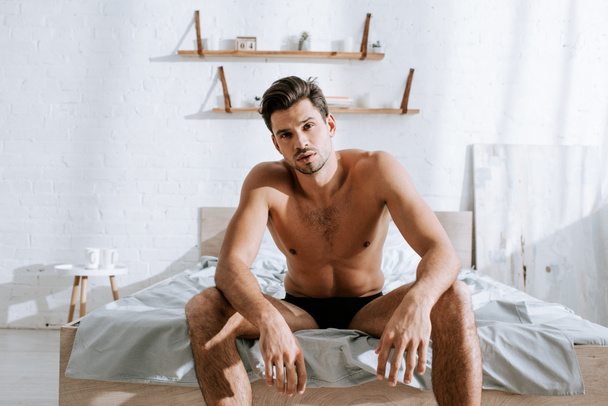 bez koszulki mężczyzna patrząc w aparat podczas siedzenia na łóżku  - Zdjęcie, obraz