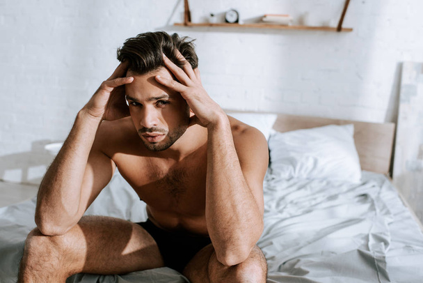 shirtless man looking at camera while sitting on bed and touching head  - Фото, зображення