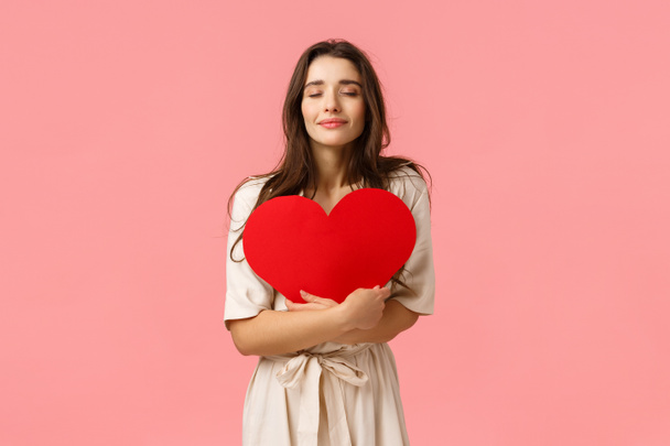 Care, tenderness and beauty concept. Tender and lovely young girlfriend in dress standing over pink background, hugging large heart card with closed eyes and cute smile, standing pink background - Foto, afbeelding