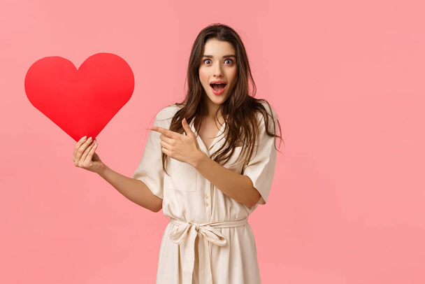 Amazement, tenderness and romance concept. Attractive surprised and intrigued young girl wonder who sent her valentines card, holding big red heart, pointing finger and gazing impressed camera - Fotografie, Obrázek