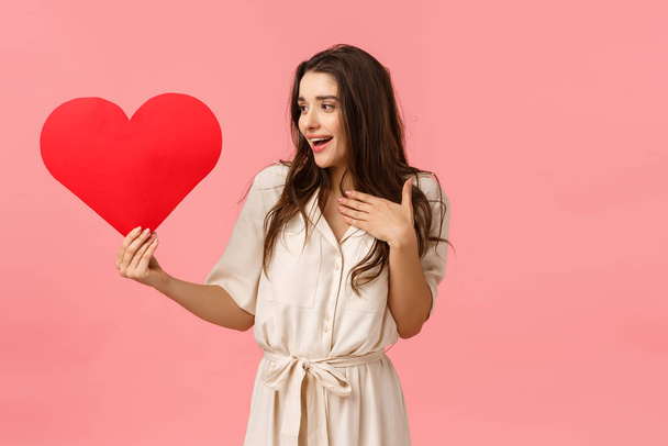 Affection, love and romance concept. Happy and alluring, tender young woman looking touched at large cute heart card, smiling pleased and surprised, touching chest grateful, pink background - Fotografie, Obrázek