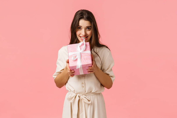 Anticipation, celebration and holidays concept. Lovely young woman in stylish light dress, biting gift box and smiling, wanting open, tempting see whats inside surprise present, pink background - Foto, afbeelding