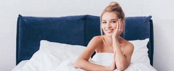 panoramic shot of beautiful smiling woman in sheets sitting on bed and looking at camera - Photo, Image
