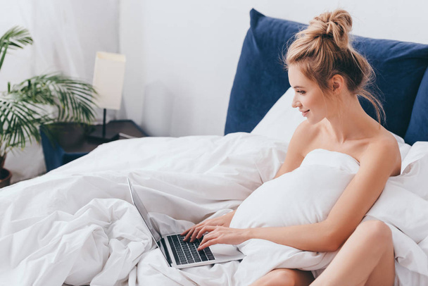 smiling woman using laptop on bed with sheets in morning  - Zdjęcie, obraz
