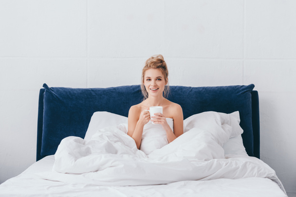 усміхнена жінка з чашкою кави сидить у ліжку вранці
  - Фото, зображення