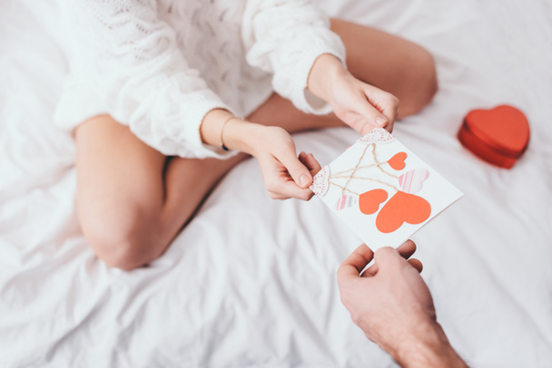 vista recortada de la mujer tomando tarjeta de felicitación con corazones para el día de San Valentín del hombre en la cama con caja de regalo
 - Foto, Imagen
