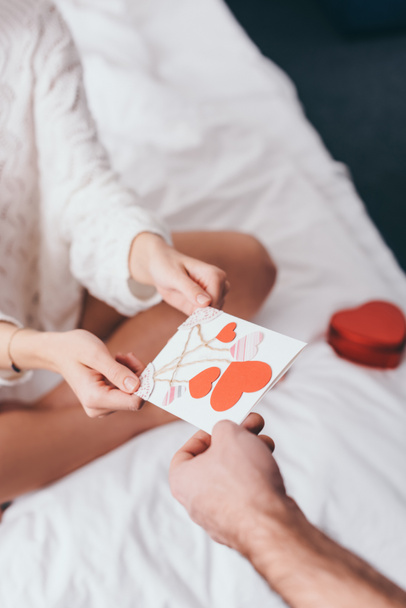 vista recortada de pareja con tarjeta de felicitación y caja de regalo para el día de San Valentín en la cama
 - Foto, Imagen