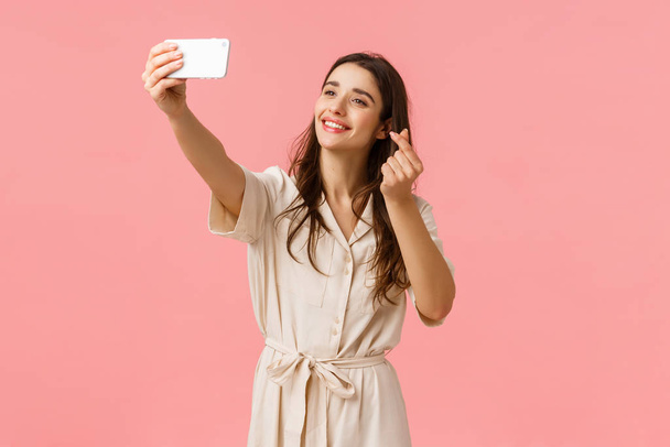 Wesoły piękny młody Europejczyk kobieta za pomocą filtra do robienia cute selfie, fotografowania na aparacie komórkowym, uśmiechając się i pokazując koreański gest serca na smartfonie, stojące różowe tło - Zdjęcie, obraz