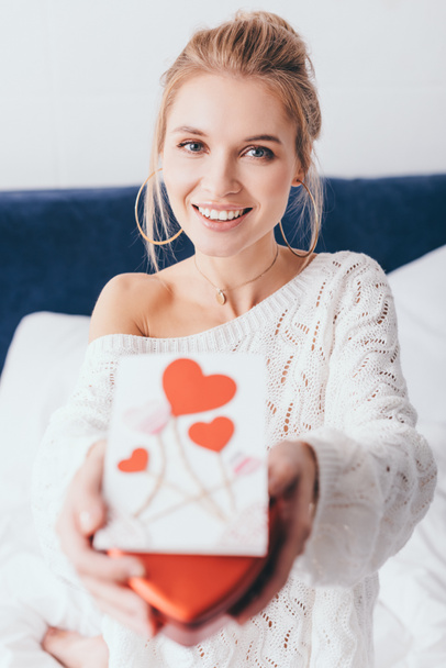 selective focus of happy woman holding gift box and valentine card on bed  - Fotoğraf, Görsel
