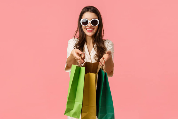 Шопінг, концепція споживачів. Весела приваблива брюнетка, тримає сумки для покупок і посміхається, одягнена в окуляри, насолоджується переглядом через магазини, стоїть рожевий фон радісний
 - Фото, зображення