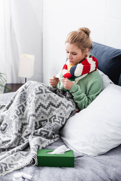 upset sick woman in scarf holding pills in bed with napkins - Fotó, kép