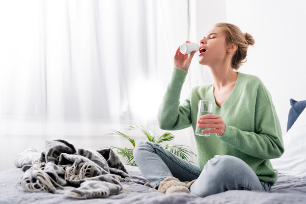 mujer enferma tomando pastillas de la botella y sosteniendo un vaso de agua en el dormitorio
 - Foto, Imagen